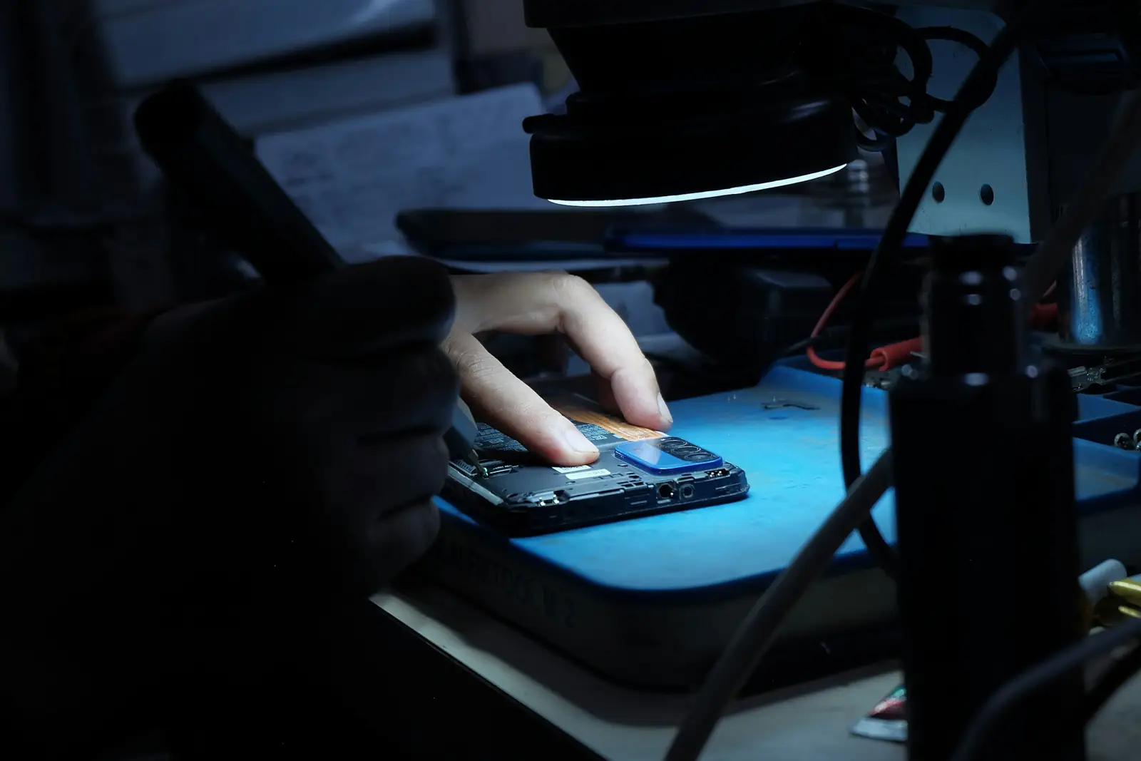 Des mains gantées travaillent sur un appareil ouvert, sous une lampe d’atelier, dans la pénombre.