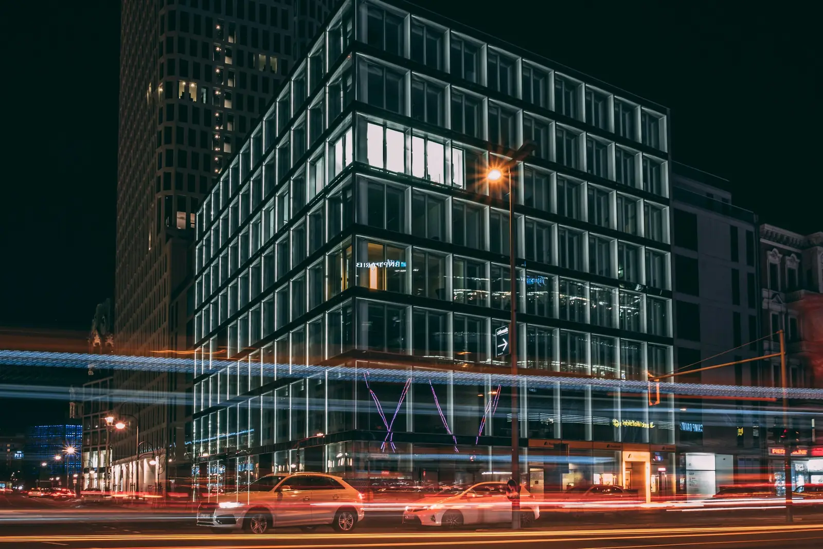 Un immeuble de bureaux la nuit, vu depuis le carrefour, les traînées des phares au premier plan.