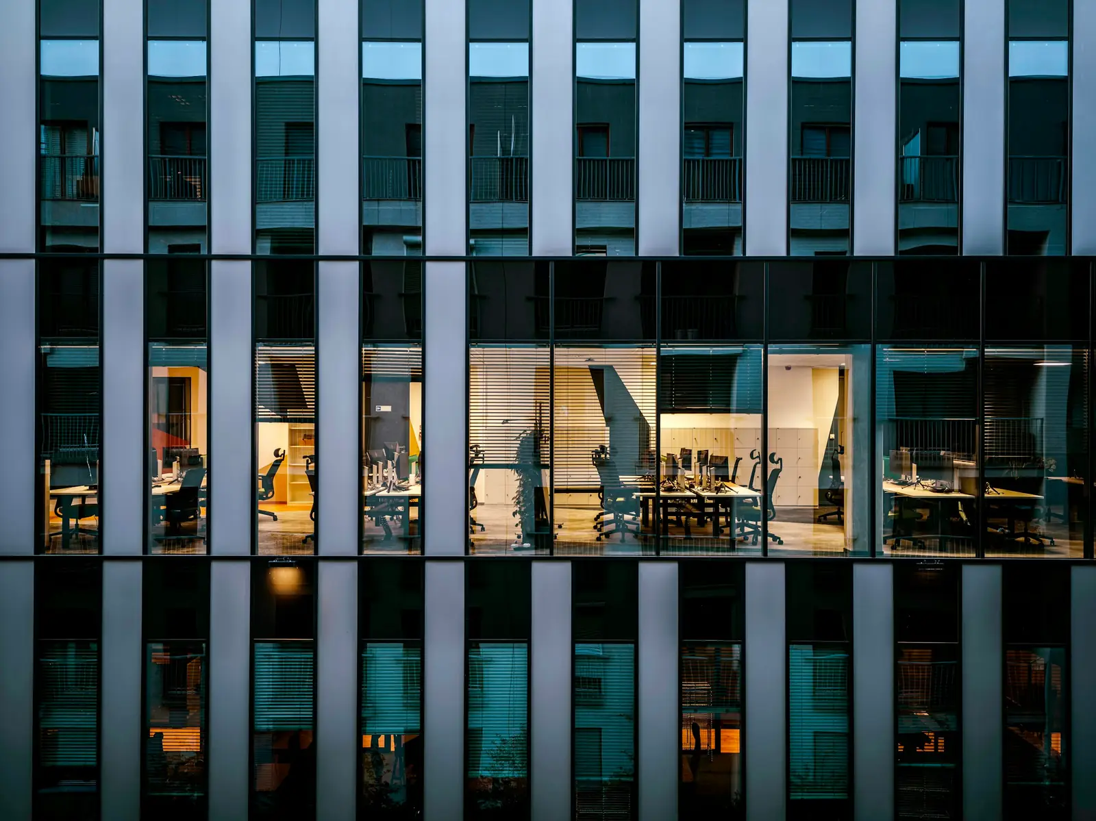 Un immeuble de bureaux la nuit, vu à travers des lames verticales : les plateaux éclairés derrière les vitres.