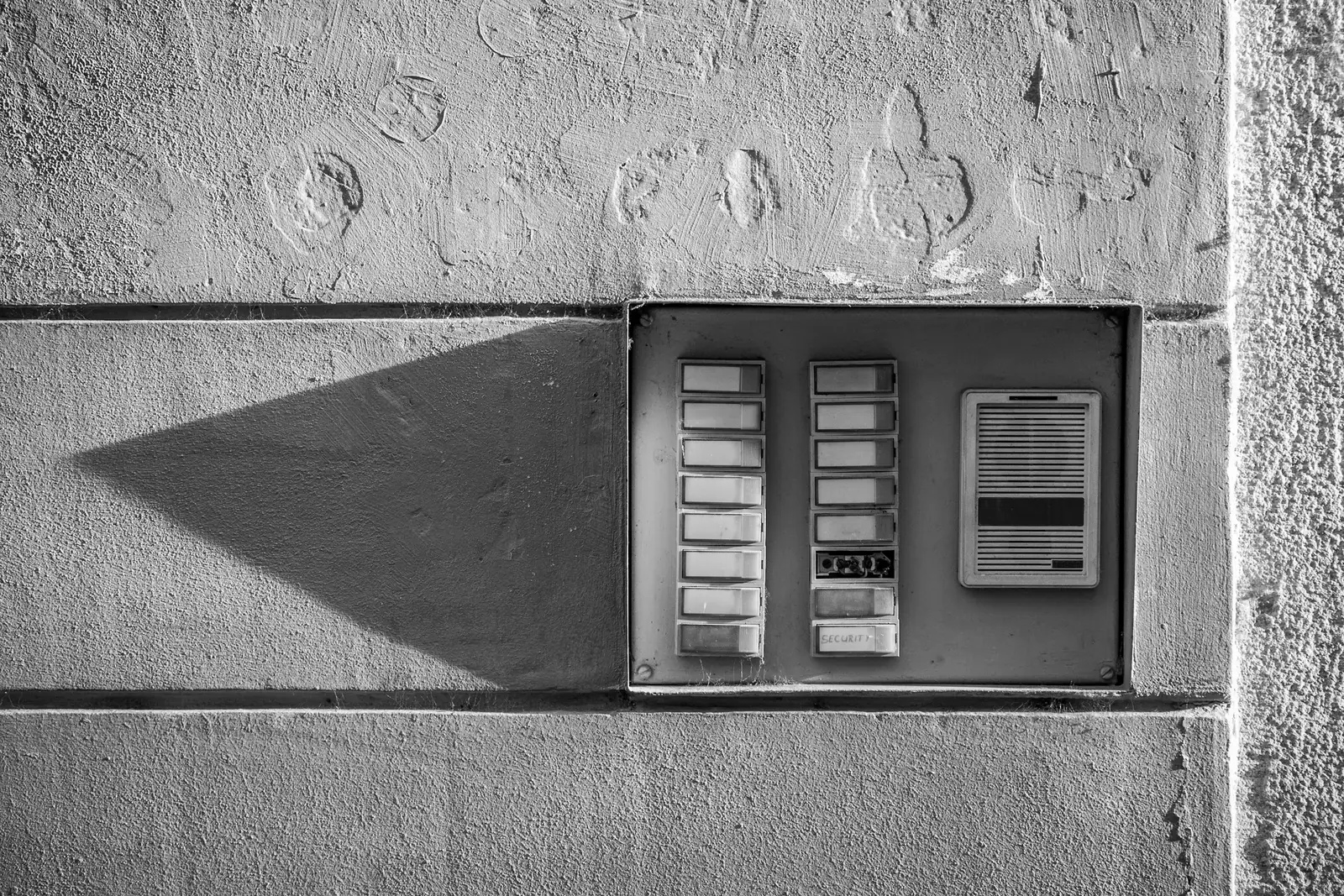Un panneau d’interphone encastré dans un mur de béton, dans une lumière rasante — photographie en noir et blanc.