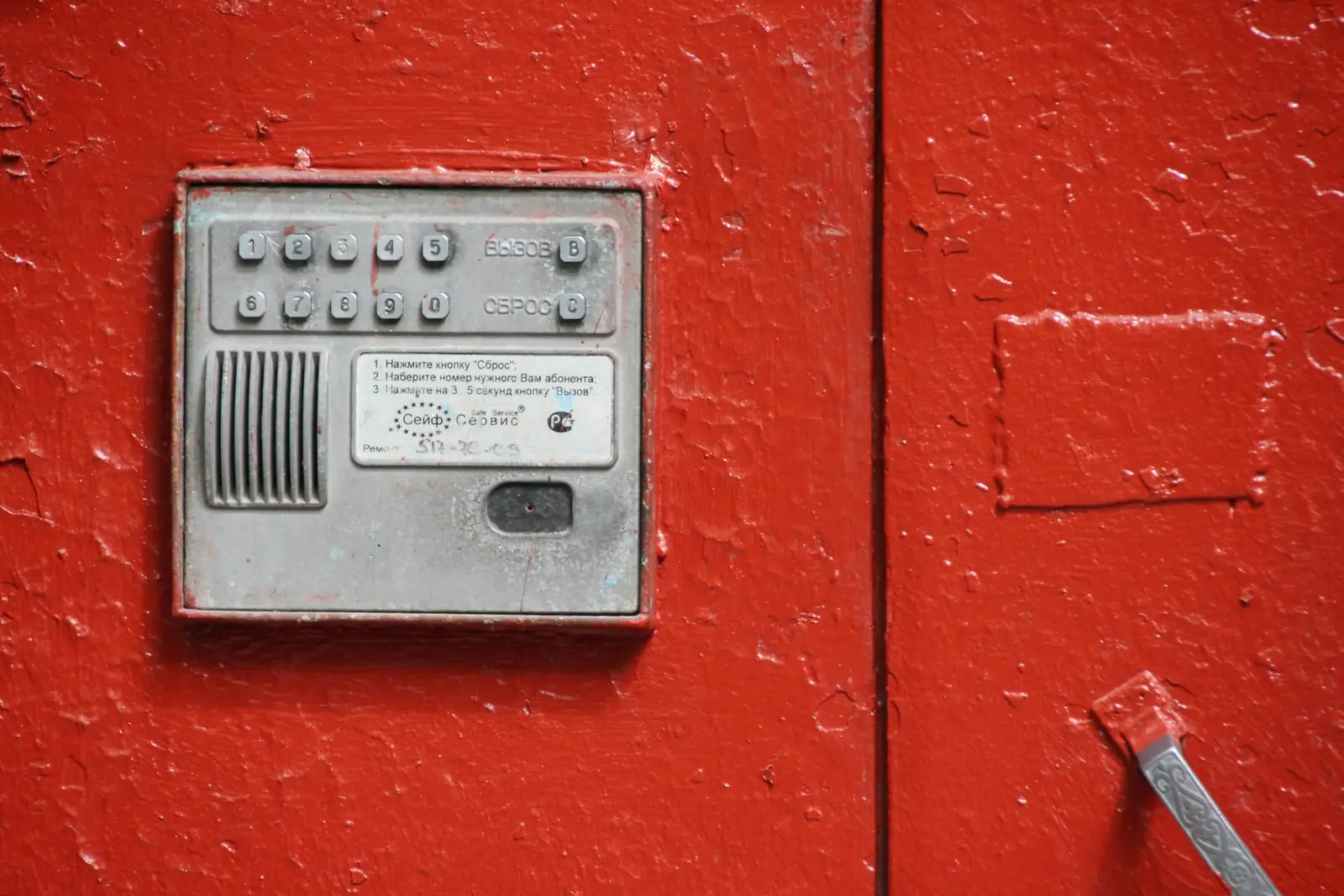 Un interphone d’immeuble en métal brossé, vissé sur une porte peinte en rouge vif.