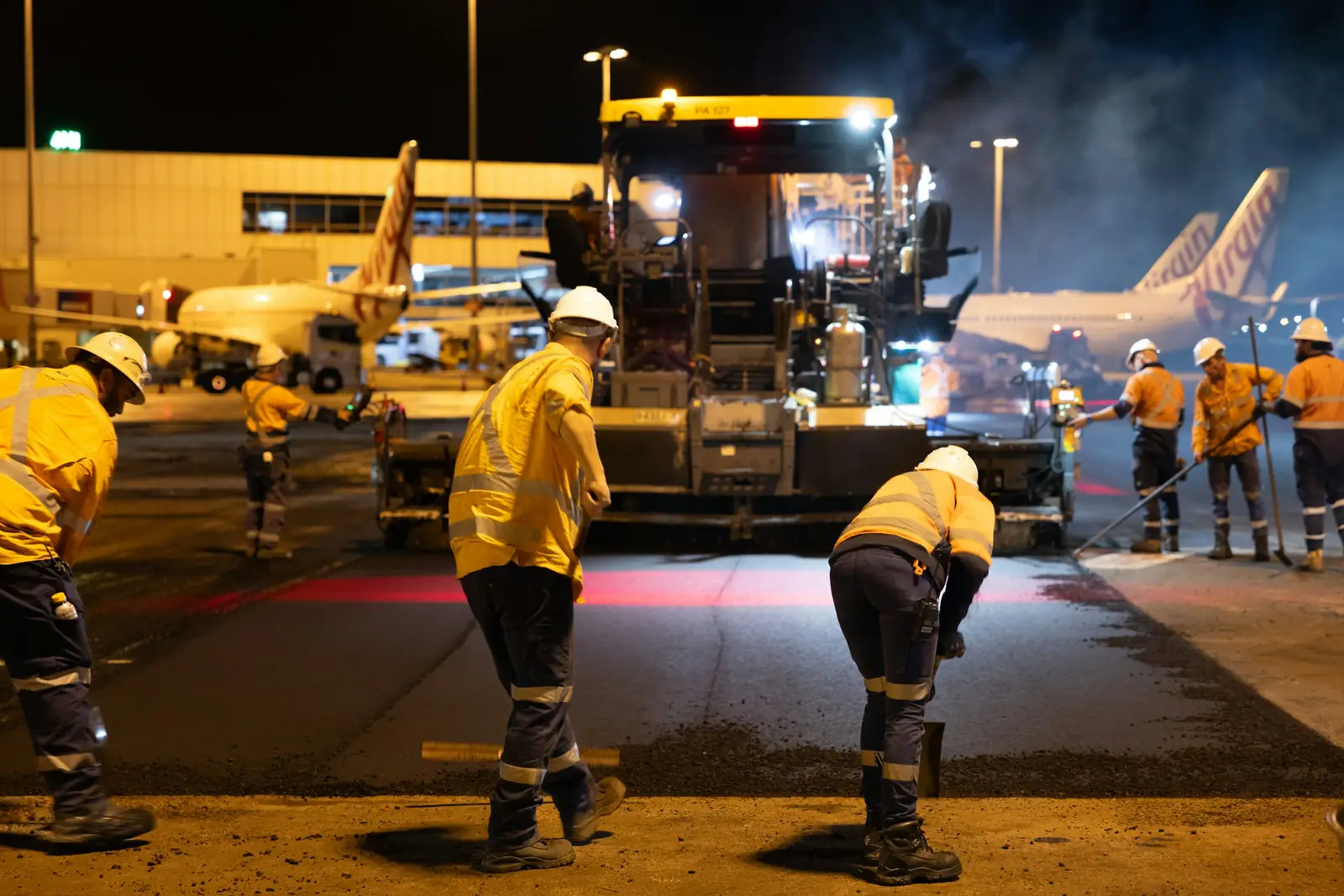 Un chantier de nuit sous les projecteurs : plusieurs personnes en tenue haute visibilité autour d’un engin, la chaussée ouverte.