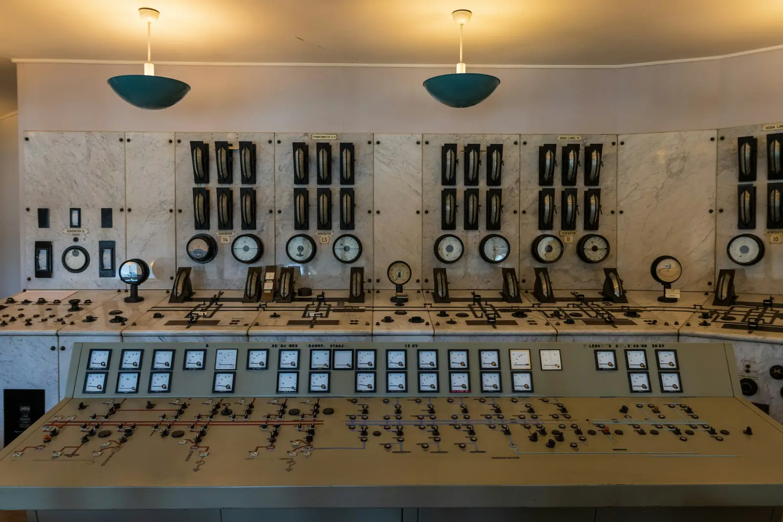Un pupitre de commande ancien dans une salle de contrôle : deux rangées de cadrans, des interrupteurs alignés et deux lampes suspendues au-dessus.