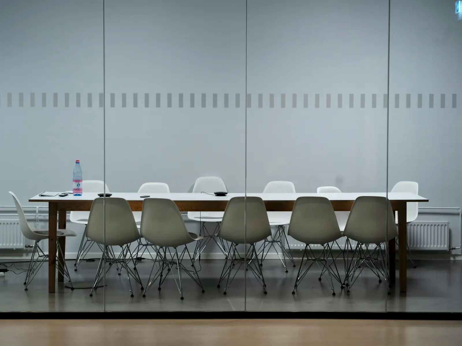 Une salle de réunion vitrée, vide : une longue table blanche, des chaises alignées et la lumière du jour sur toute la cloison.